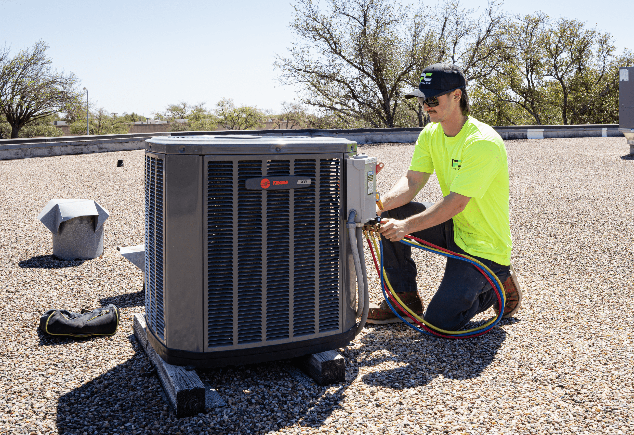A technician in a neon yellow shirt checks a rooftop HVAC unit with gauges and hoses on a gravel surface under a clear sky.